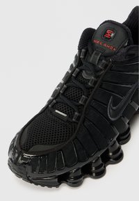 Fekete Nike Shox sportcipők hálós felsőrésszel és szintetikus túllépésekkel rendelkeznek, lekerekített orrukkal, megemelt talpukkal, amely egyedi gumioszlopokkal van ellátva, és szürke akcentussal.