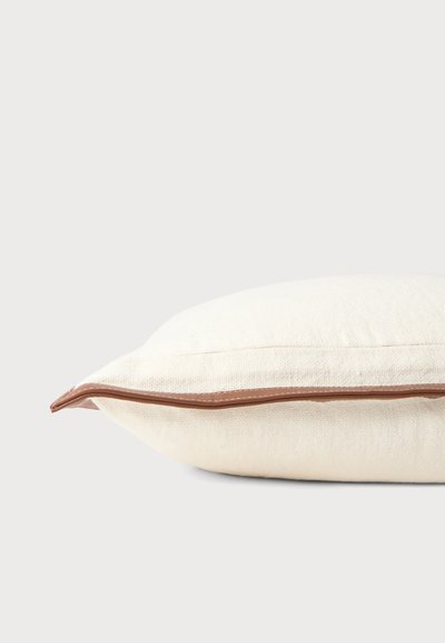 Coussin en tissu couleur crème avec une fermeture éclair marron le long d'un bord sur un fond gris clair.
