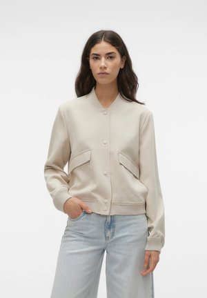 Jeune femme aux longs cheveux foncés porte une veste bomber beige boutonnée et un jean bleu clair, debout avec une main dans la poche.