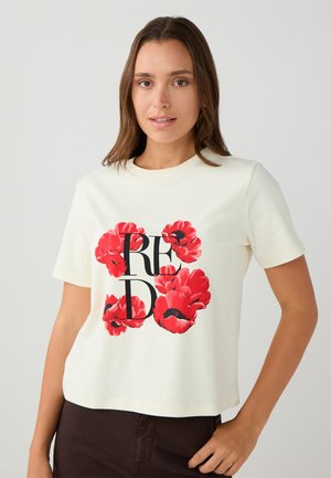 Jonge vrouw met bruin haar draagt een wit T-shirt met een rode bloemenprint en zwarte letters "RED," gecombineerd met een donkere broek.