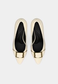 Pointed-toe heels in cream leather with a gold buckle detail. Interior features a black cushioned insole for comfort. Smooth texture throughout.