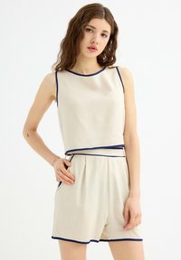 Conjunto de dos piezas beige que incluye un top corto sin mangas con ribete azul marino y pantalones cortos de talle alto a juego con bolsillos laterales y plisados.