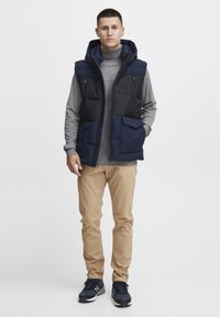 Padded vest in navy and black, featuring a hood, two front pockets, and a zip closure. Worn over a gray turtleneck and paired with tan pants.