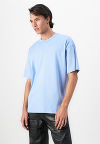 Young man wearing a light blue oversized T-shirt and black leather cargo pants, standing against a white background.