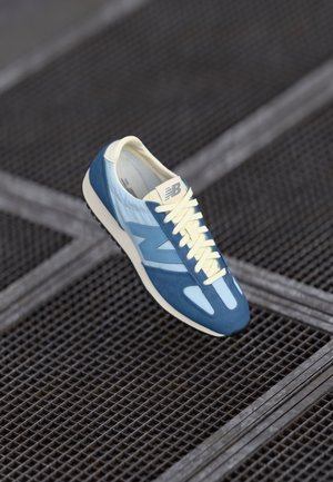 Niebiesko-jasnoniebieski sneaker New Balance z kremowymi sznurowadłami, ustawiony pod kątem na metalowej podłodze z krat.