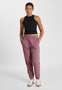 Débardeur noir côtelé et pantalon de jogging rose clair avec une taille élastique. Baskets blanches avec semelle en caoutchouc. Le modèle se tient devant un fond blanc.