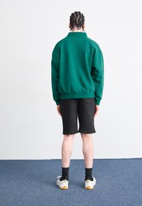 Sweat-shirt vert foncé à coupe décontractée et poignets côtelés, associé à un short noir. Baskets à semelle en caoutchouc visibles en dessous.