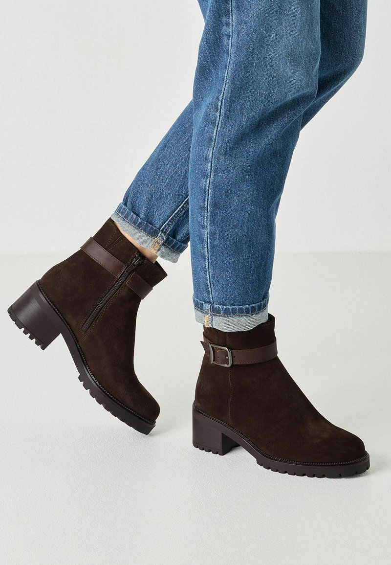 Personne portant un jean bleu et des bottines en daim marron foncé avec talons carrés et fermetures éclair latérales, debout sur un sol clair.