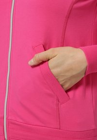 Virkistävän pinkki vetoketjullinen takki, valmistettu pehmeästä kankaasta, jossa on sivutasku ja resorit hihansuissa ja helmassa.