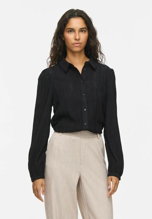 Femme aux cheveux bouclés foncés portant un chemisier noir à manches longues boutonné et un pantalon beige clair taille haute, debout devant un fond uni.