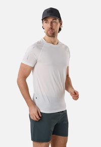 Homme portant une casquette noire, un t-shirt de sport blanc à manches courtes et un short gris foncé, debout sur un fond neutre.