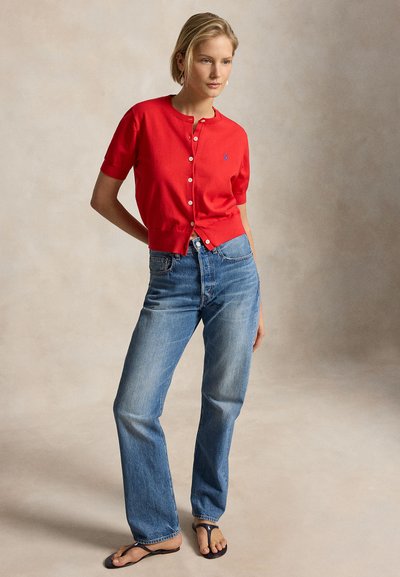 Nő piros, rövid ujjú gombos kardigánban, kék, magas derekú farmerben és fekete papucsban, semleges háttér előtt áll.