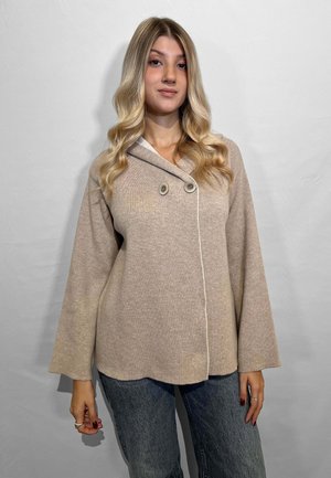 Cardigan lavorato a maglia beige con collo a scialle, dotato di due grandi bottoni e rifiniture bianche, abbinato a pantaloni in denim blu.