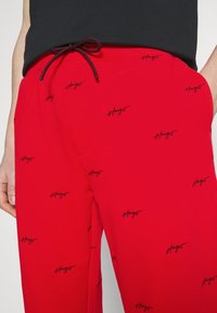 Rote Jogginghosen mit einem schwarzen, stickereiartigen Logomuster, elastischem Bund mit Kordelzug und Seitentaschen. Glattes, lässiges Material.
