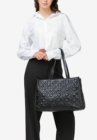 Borsa tote in pelle nera con motivo trapuntato geometrico, dotata di due manici lunghi. Modello che indossa una camicia bianca e pantaloni neri.