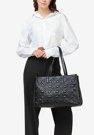 Borsa tote in pelle nera con motivo trapuntato geometrico, dotata di due manici lunghi. Modello che indossa una camicia bianca e pantaloni neri.