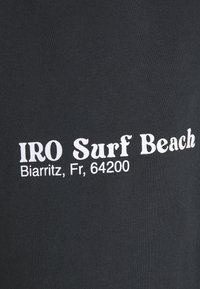 Czarna bawełniana koszulka z białym napisem "IRO Surf Beach" oraz lokalizacją "Biarritz, Fr, 64200." Gładka faktura, casualowy design.