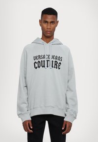 Ανοιχτό γκρι φούτερ με μπροστινή τσέπη καγκουρό, κουκούλα με κορδόνι και μαύρο κείμενο "VERSACE JEANS COUTURE." Διαθέτει ριμπ μανσέτες και τελειώμα.