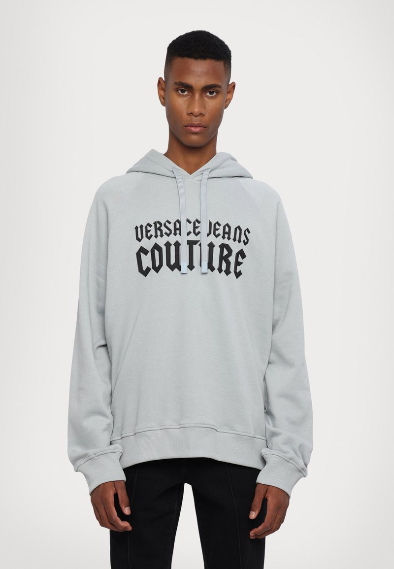 Ανοιχτό γκρι φούτερ με μπροστινή τσέπη καγκουρό, κουκούλα με κορδόνι και μαύρο κείμενο "VERSACE JEANS COUTURE." Διαθέτει ριμπ μανσέτες και τελειώμα.