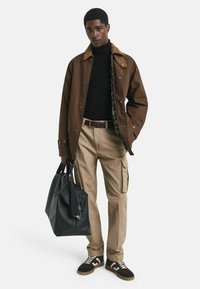 Junger Mann in brauner Jacke über einem schwarzen Rollkragenpullover, beigen Cargo-Hosen und schwarzen Turnschuhen, der eine große schwarze Reisetasche hält und steht.