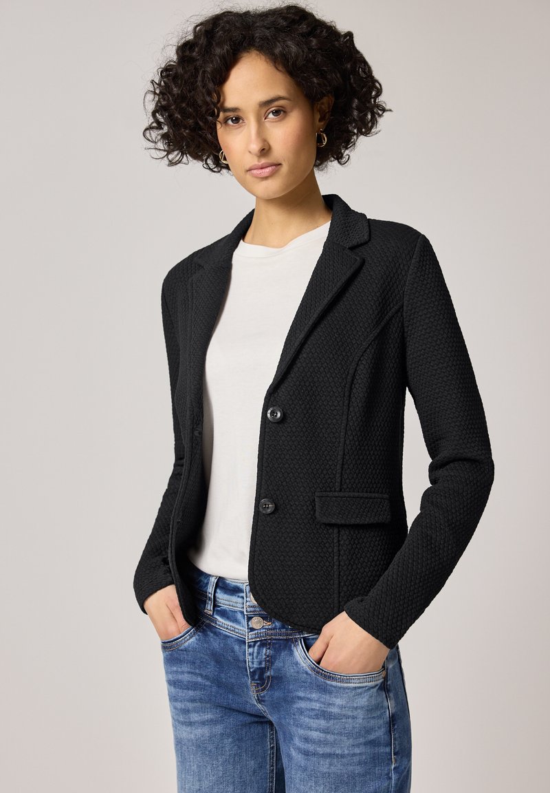 Femme aux cheveux bouclés portant un blazer noir texturé sur une chemise blanche et un jean bleu, mains dans les poches, sur un fond clair uni.