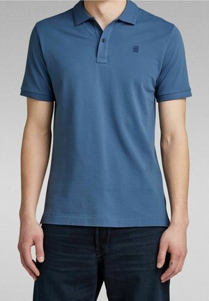 Mężczyzna w prostym niebieskim polo z krótkim rękawem, z małym logo na piersi, oraz ciemnoniebieskich dżinsach, stojący na jednolitym tle.