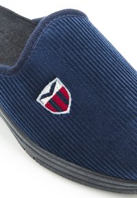 Pantuflas de corduroy en color azul marino con suela de goma negra. Presentan un logotipo bordado de un escudo en rojo, blanco y azul en el lateral.