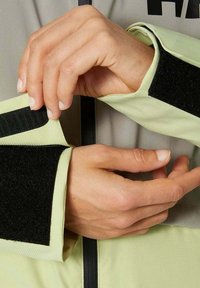 Hellgrüne Jacke aus strukturiertem Stoff, mit schwarzen Klettverschlussmanschetten und einem Reißverschluss. Nahaufnahme von Händen, die den Ärmel anpassen.