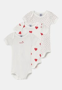 Drei weiße Babykörperanzüge aus Baumwollstoff. Einer mit roten Herzen, einer mit kleinen roten Punkten und einer mit dem Schriftzug "Petit Bateau" in roter Schrift.