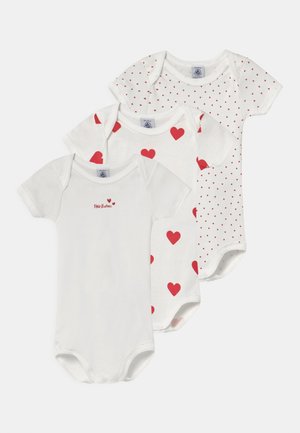 Trois bodies pour bébés blancs en coton. L'un avec des cœurs rouges, un autre avec de petits pois rouges et un dernier arborant "Petit Bateau" en écriture rouge.