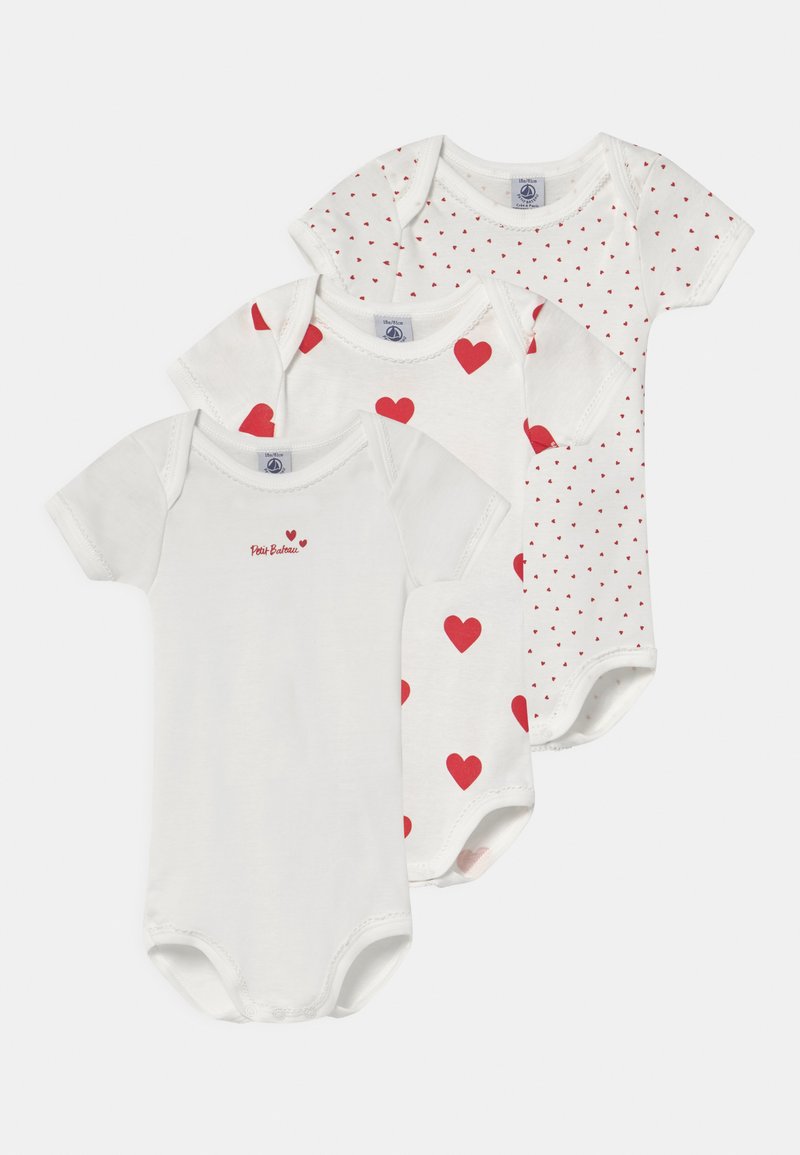 Drei weiße Babykörperanzüge aus Baumwollstoff. Einer mit roten Herzen, einer mit kleinen roten Punkten und einer mit dem Schriftzug "Petit Bateau" in roter Schrift.