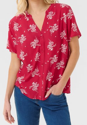 Femme portant un chemisier rouge à manches courtes avec un motif floral blanc et un jean bleu, une main dans la poche, sur un fond uni.