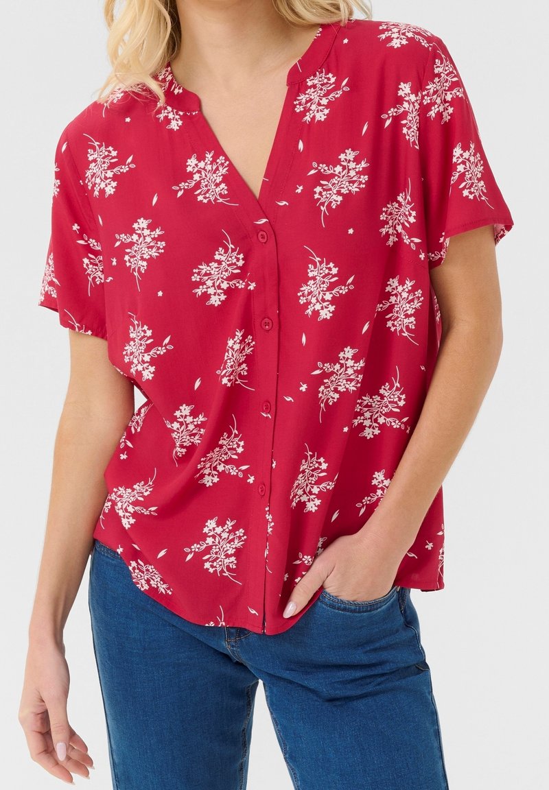 Kvinde iført rød kortærmet skjorte med knapper og hvidt blomsterprint samt blå jeans, den ene hånd i lommen, foran ensfarvet baggrund.