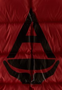 Rød pufferjakke med et stort sort trekantet logo og håndgrafik i midten. Stoffet har en glat, skinne overflade.