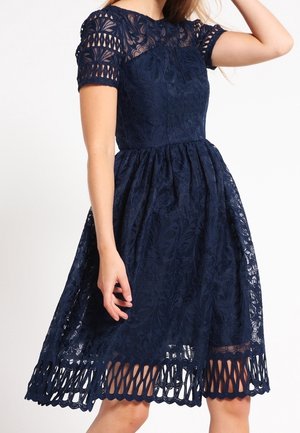 Donna che indossa un vestito di pizzo blu navy al ginocchio con maniche corte e orlo smerlato, in piedi davanti a uno sfondo semplice.