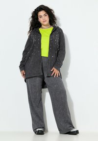 Chaqueta gris con cremallera y acentos de estrella plateada, superpuesta a una camiseta verde brillante y pantalones anchos grises a juego, combinada con zapatos negros brillantes.