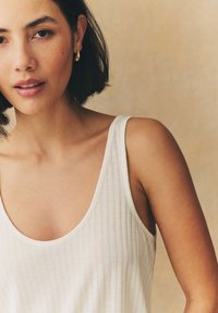 Débardeur blanc côtelé avec un large décolleté et des fines bretelles, mettant en valeur une texture lisse. Des boucles d'oreilles pendantes dorées accentuent le design.