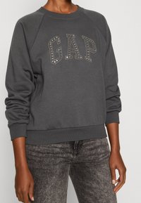 Felpa grigia con maniche raglan e polsini a costine, caratterizzata da una scritta decorativa "GAP" in strass sulla parte anteriore. Texture morbida, vestibilità casual.
