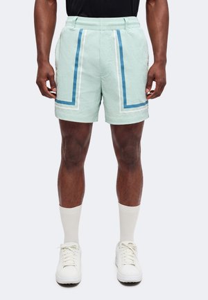 Mand iført lysegrønne shorts med blå og hvide geometriske striber, hvide ankelsokker, hvide sneakers og en sort top, stående oprejst.