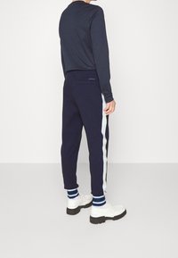 Námořnické joggery s světle modrým pruhem na boku, spárované s bílými botami a pruhovanými ponožkami. Obsahují detail zadní kapsy.