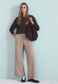 Femme debout contre un mur bleu pâle portant un pull marron, un pantalon large beige, des baskets noires, tenant un sac à main marron assorti.