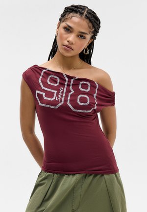 Νεαρή γυναίκα με πλεξούδες, φορούντος μπορντό τοπ με ακάλυπτους ώμους με αριθμό 98 και ελιάς παντελόνι, ποζάρει σε απλό φόντο.