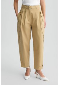 Pantalons cargo beiges avec poches latérales, taille haute et une ceinture avec une boucle métallique ronde. Des boutons plats soulignent les poches.