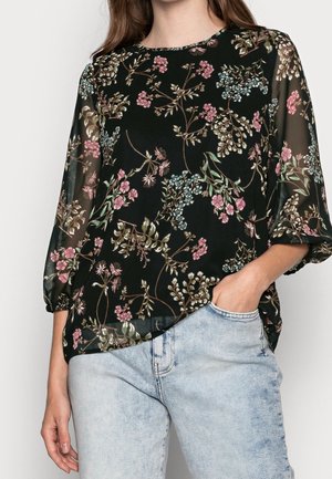 Vrouw die een zwarte doorschijnende blouse draagt met een roze, blauw en groen bloemenpatroon en lichtblauwe spijkerbroek, met een hand in haar zak.