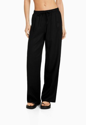 Kobieta w luźnych czarnych spodniach z troczkiem i czarnych sandałach, stojąca na białym tle.