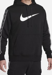 Person trägt schwarzen Hoodie mit weißem Nike-Swoosh-Logo auf der Brust und passende schwarze Hose mit gestreiften Details an Ärmeln und Beinen.