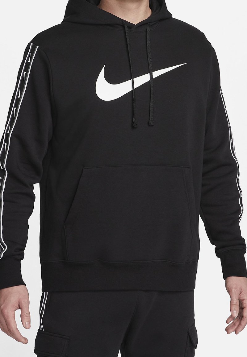 Person trägt schwarzen Hoodie mit weißem Nike-Swoosh-Logo auf der Brust und passende schwarze Hose mit gestreiften Details an Ärmeln und Beinen.