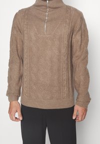 Beiger Strickpullover mit hohem Kragen und halbem Reißverschluss, mit strukturierten Zopfmustern sowie gerippten Bündchen und Saum.