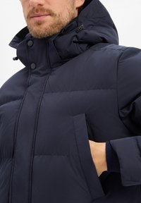 Marineblaue Steppjacke mit hohem Kragen, Kapuze mit Reißverschluss und großer aufgesetzter Tasche. Glatte Oberfläche und gesteppte Optik. Verstecktes Bündchen-Detail.