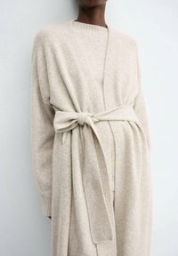 Cardigan lavorato a maglia beige realizzato in materiale morbido, con cintura a fiocco e maniche lunghe, dalla vestibilità ampia e colletto rotondo.